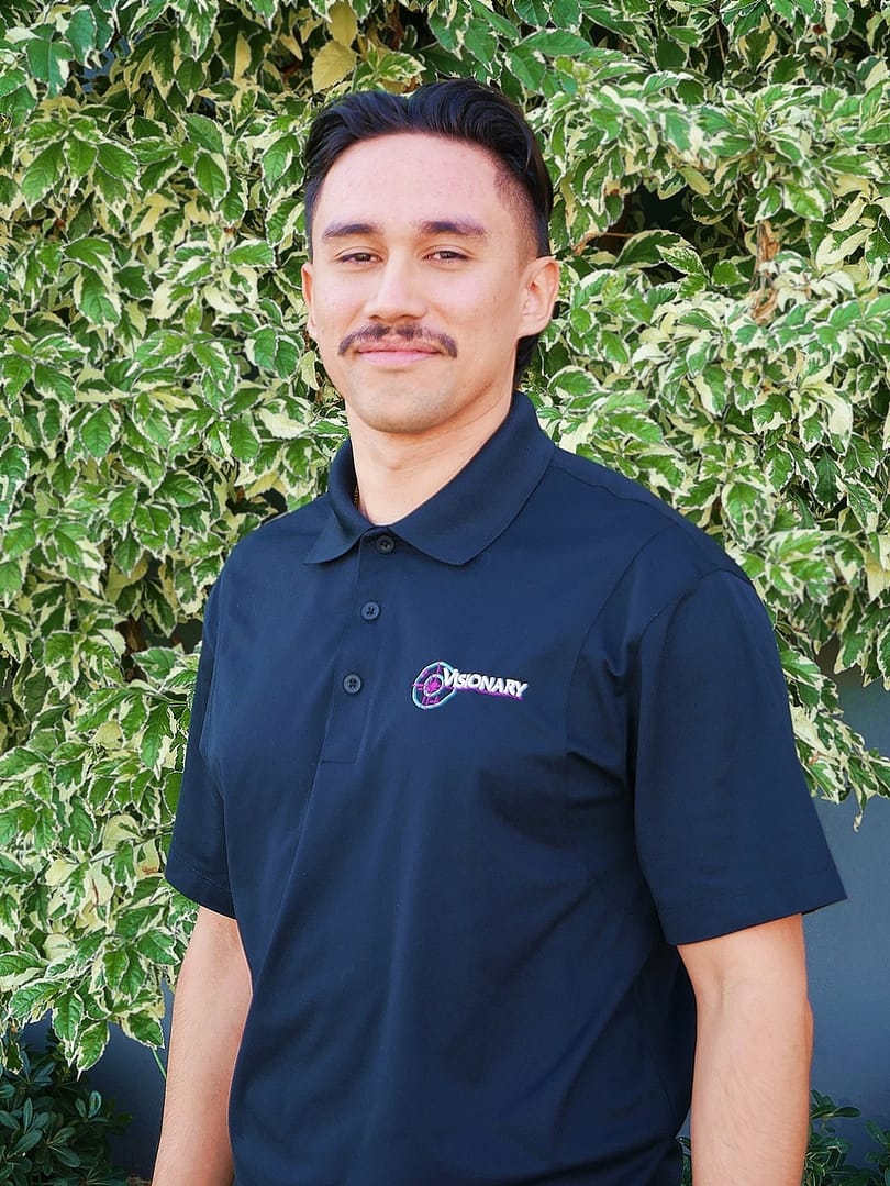 Smiling man with black hair standing in front of a grey wall and a green bush.