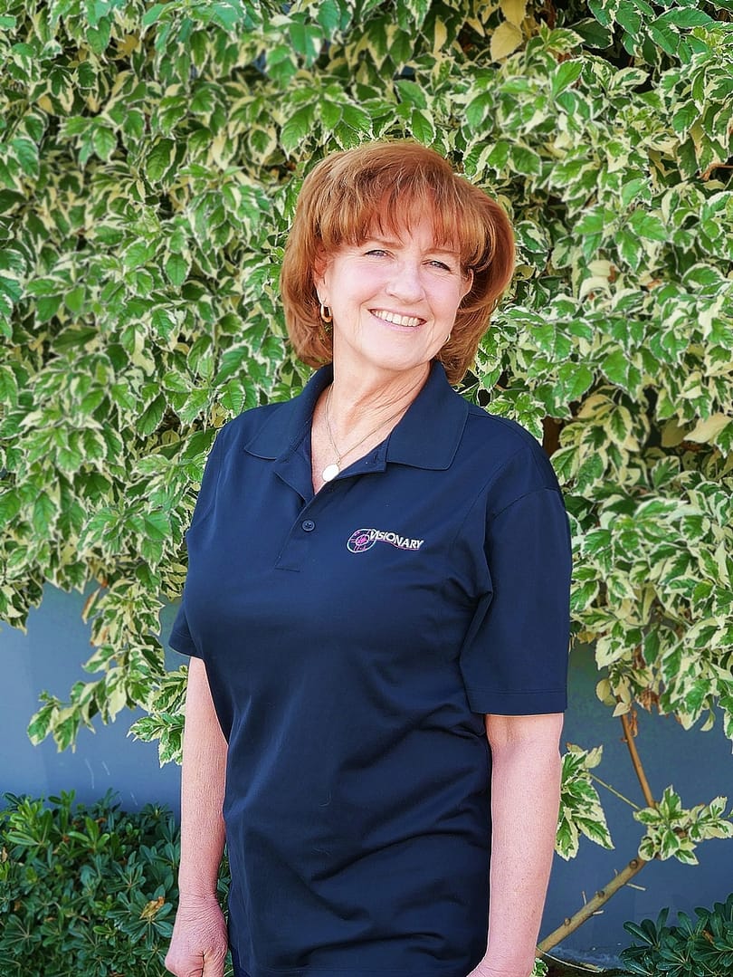 Smiling women with red hair standing in front of a green bush and a grey wall.