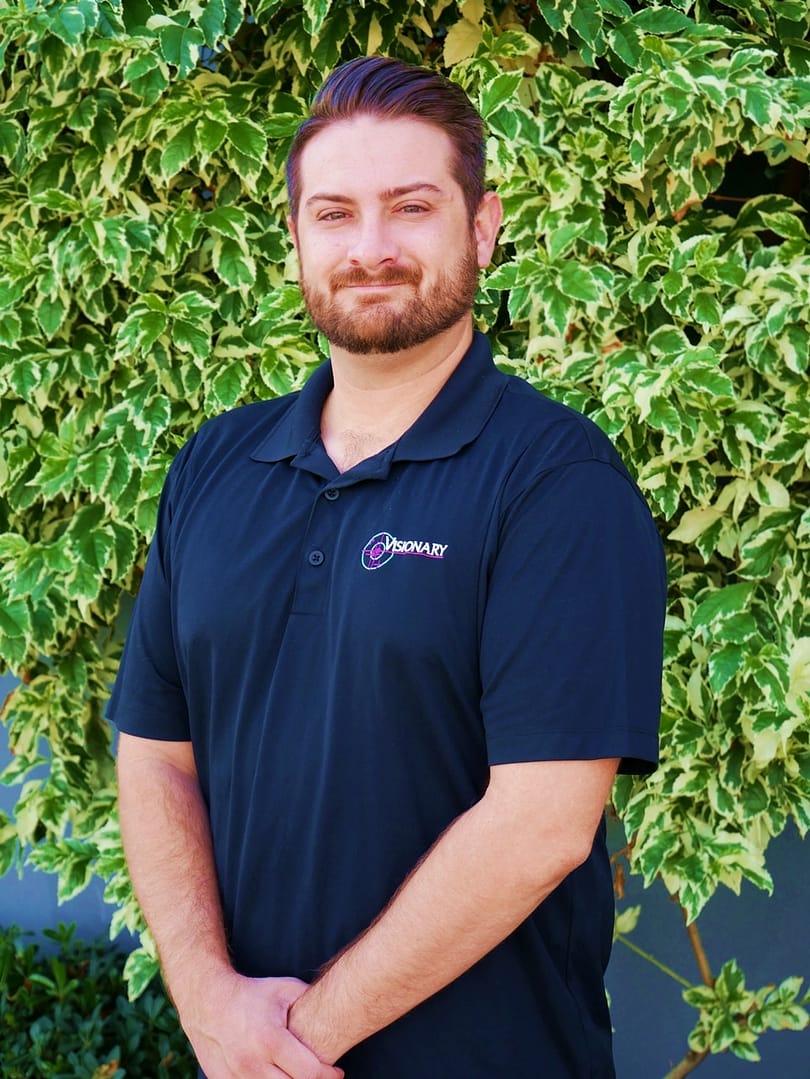 Smiling brown haired man standing in front of a green bush and grey wall.