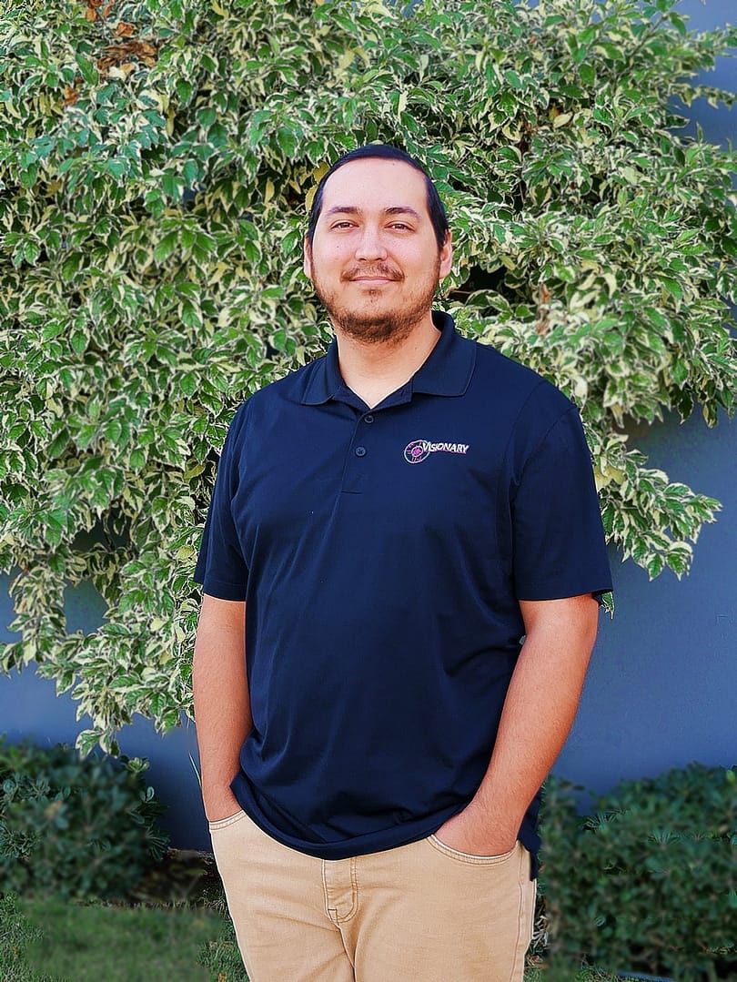 Smiling man standing in front of a grey wall and green tree.